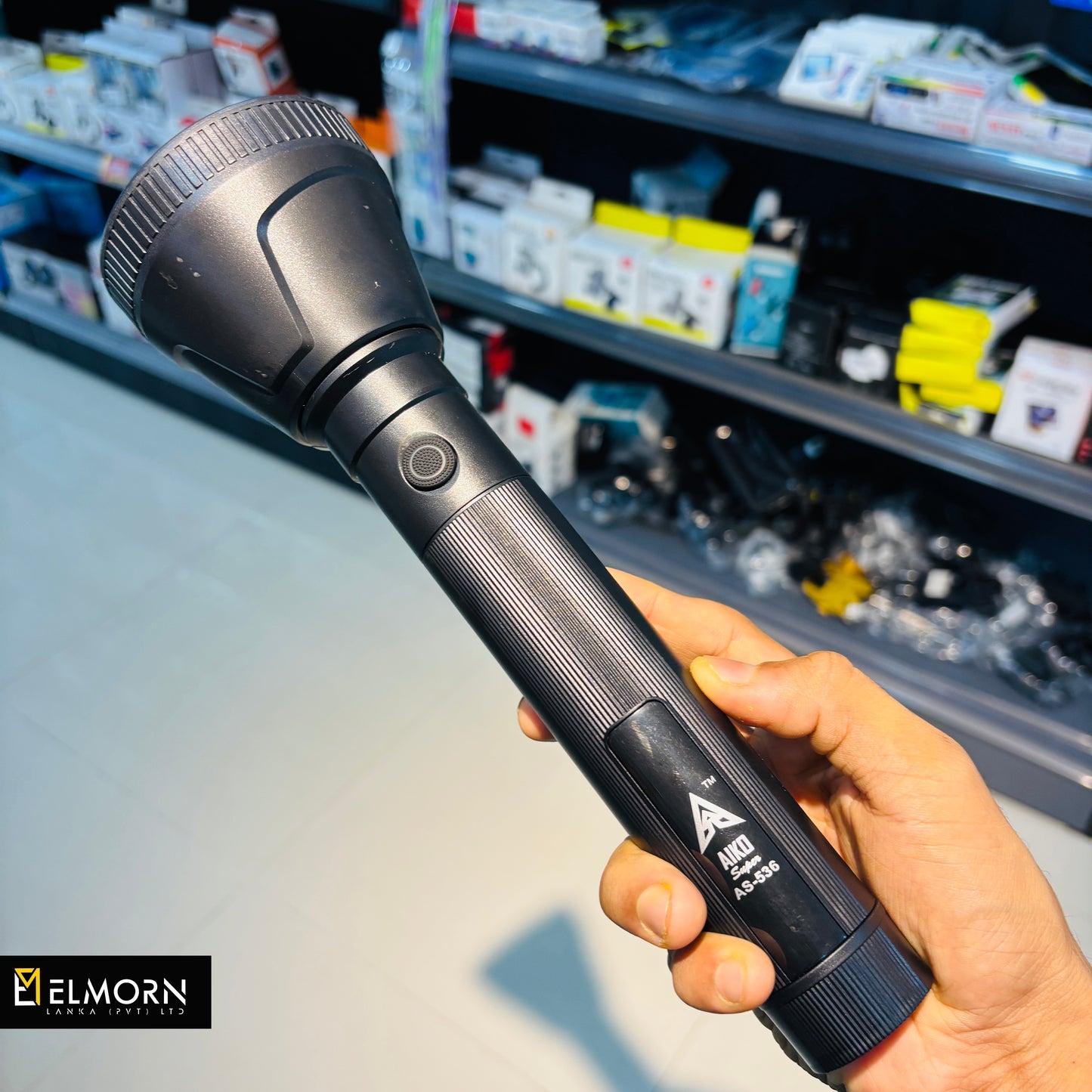 A person's hand holding a black Elmorn As 536 flashlight in a store setting with shelves of products blurred in the background.