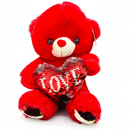 A red plush teddy bear holding a sequined heart-shaped pillow with the word 'LOVE' in white, photographed against a plain white background.