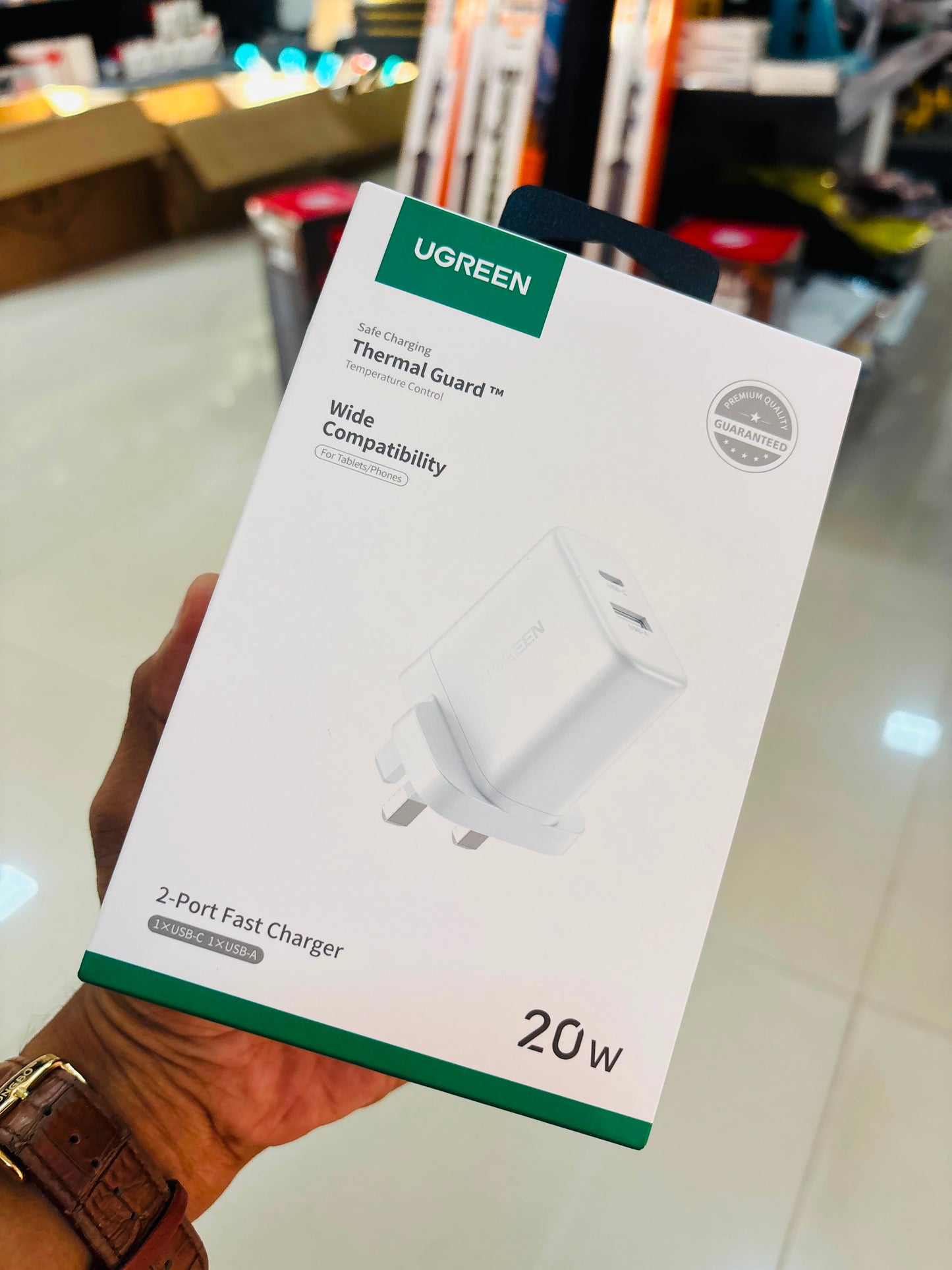 A hand holding a UGREEN 20W thermal guard 2-port fast charger box in a retail store setting.